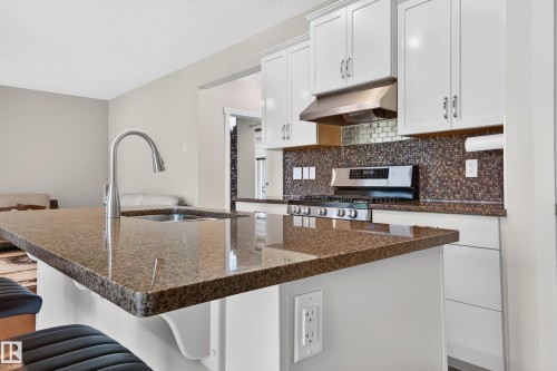 Kitchen featuring an island with a built-in sink and faucet, brown speckled countertops, white upper and lower cabinetry, a mosaic tile backsplash, and a stainless steel gas range with a matching range hood - 1512 Cunningham Cape, Edmonton, AB 