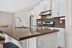 Kitchen featuring an island with a built-in sink and faucet, brown speckled countertops, white upper and lower cabinetry, a mosaic tile backsplash, and a stainless steel gas range with a matching range hood - 