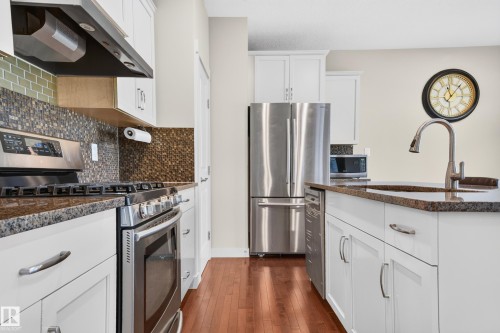 Kitchen featuring a stainless steel appliance suite, white cabinetry with silver hardware, brown granite-finish countertops, and wood-finish flooring - 1512 Cunningham Cape, Edmonton, AB 