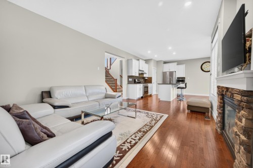 Open-concept living space featuring wood-finish flooring, a stone-front fireplace with a white mantle, recessed lighting, white kitchen cabinetry, and stainless steel appliances - 1512 Cunningham Cape, Edmonton, AB 