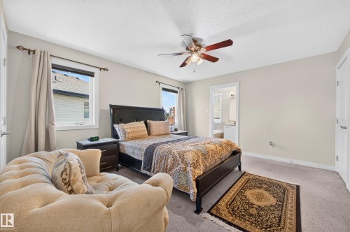 Carpeted room featuring a ceiling fan with integrated lighting, two windows with white trim, and a doorway opening to a bathroom with a white vanity - 1512 Cunningham Cape, Edmonton, AB 