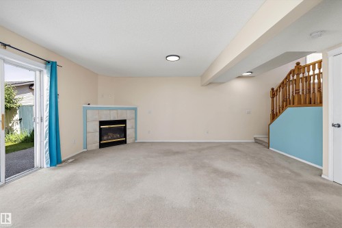 Carpeted lower-level living area featuring a corner fireplace with a tile surround, a sliding glass patio door, and a wooden staircase with a painted knee wall - 15016 134 Street, Edmonton, AB 