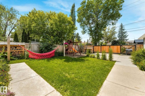 Expansive rear yard featuring a lush lawn, concrete pathways, a wooden privacy fence, a wood storage rack, and a swing set - 8212 148 Street Nw, Edmonton, AB 
