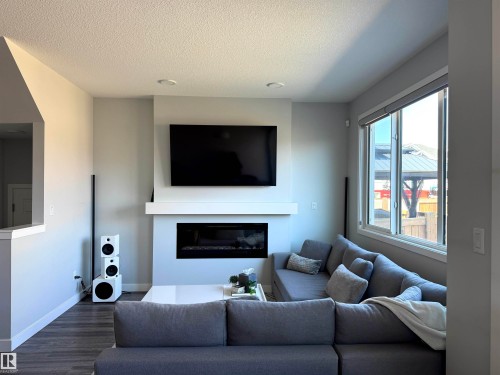 Living area featuring a contemporary fireplace with a white mantel, recessed lighting, wood-finish flooring, and a large window - 7432 Elmer Road, Edmonton, AB 