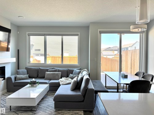 Open concept living space featuring a contemporary fireplace with a white mantel, large windows with roller shades, and a sliding glass door leading to an outdoor deck - 7432 Elmer Road, Edmonton, AB 