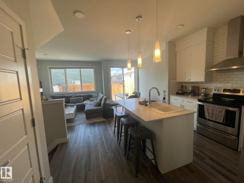 Open-concept living space featuring a kitchen island with dual sinks, pendant lighting, stainless steel appliances, and wood-finish flooring - 7432 Elmer Road, Edmonton, AB 