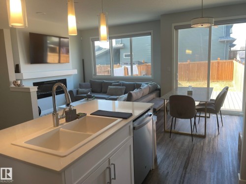 Kitchen island with a double basin sink and stainless steel pull-down faucet, open to a living area featuring an electric fireplace, and a dining space with wood-finish flooring and sliding glass door access to an outdoor deck - 7432 Elmer Road, Edmonton, AB 