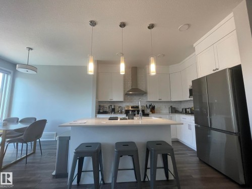 Modern kitchen featuring white cabinetry, a stainless steel refrigerator, and a kitchen island with pendant lighting - 7432 Elmer Road, Edmonton, AB 