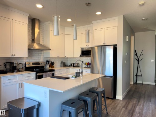 Modern kitchen featuring white shaker cabinetry, stainless steel appliances, a subway tile backsplash, a central island with bar seating, and recessed and pendant lighting - 7432 Elmer Road, Edmonton, AB 