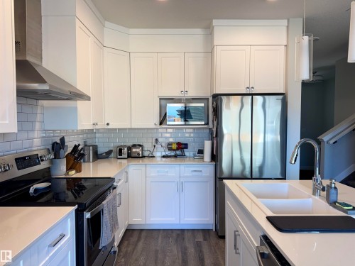 Modern kitchen featuring white shaker cabinetry, stainless steel appliances, light-toned countertops, a subway tile backsplash, and wood-finish flooring - 7432 Elmer Road, Edmonton, AB 