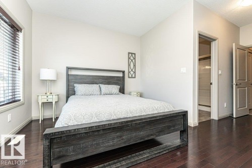 Bedroom featuring rich wood-finish flooring, light-toned walls, a window with horizontal blinds, and a doorway opening to a tiled shower enclosure - 15 175 Mcconachie Drive, Edmonton, AB 