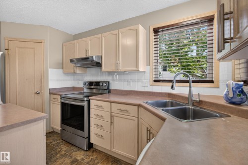 Kitchen featuring light wood-finish cabinetry, a stainless steel range with overhead ventilation, a double basin stainless steel sink with a gooseneck faucet, and a tiled backsplash - 13406 32A Street, Edmonton, AB 