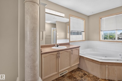 Bathroom featuring a built-in corner bathtub with white tile surround, a single vanity with a white basin sink and neutral countertop, two windows with blinds, and patterned floor tiles - 13406 32A Street, Edmonton, AB 