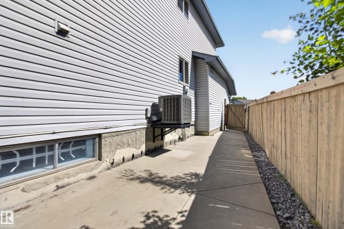 Concrete pathway alongside the property, featuring light-toned horizontal siding, a wooden privacy fence, a gate, and a visible exterior HVAC unit - 13406 32A Street, Edmonton, AB 