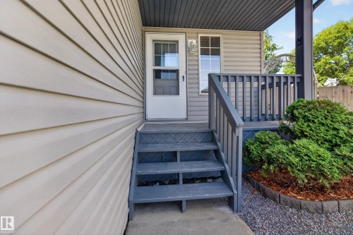 Front entry featuring a storm door with divided lites, exterior wall lantern, covered porch with dark gray railing, and steps leading to a concrete landing - 13406 32A Street, Edmonton, AB 