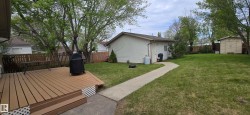 Expansive backyard featuring a large wood-finish deck with lattice skirting, a concrete pathway, a detached single-story structure with siding, a separate storage shed, and mature trees - 