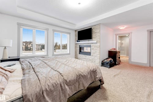 Bedroom featuring a tray ceiling, recessed fireplace with stone surround, two large windows, and an en-suite bathroom with a glass-enclosed shower - 656 180 Street Sw, Edmonton, AB - Indoor Photo Showing Bedroom