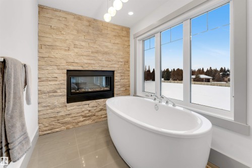 Freestanding soaking tub positioned before a large window, complemented by a floor-to-ceiling stone-faced fireplace with a linear insert - 656 180 Street Sw, Edmonton, AB - Indoor Photo Showing Bathroom With Fireplace