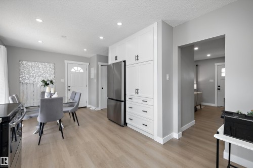 Modern kitchen and dining area featuring white cabinetry with black hardware, stainless steel refrigerator, and wood-finish flooring - 12912 103 Street, Edmonton, AB 