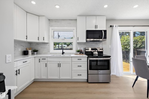 Modern kitchen featuring white shaker cabinetry, stainless steel appliances, light-toned wood-finish flooring, and a sliding glass door leading to outdoor space - 12912 103 Street, Edmonton, AB 