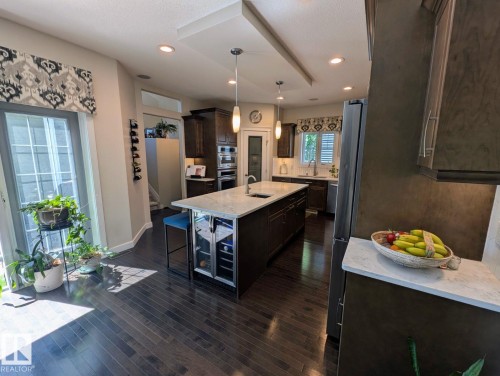 Gourmet kitchen featuring a large central island with integrated beverage cooler, dark wood-finish cabinetry, stainless steel appliances, recessed lighting, and wood-finish flooring - 2327 Frezenberg Avenue, Edmonton, AB 