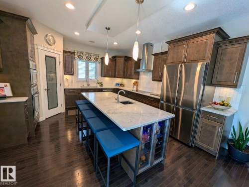 Gourmet kitchen featuring a large central island with seating, dark wood-finish flooring, white tile backsplash, stainless steel appliances, and abundant cabinetry - 2327 Frezenberg Avenue, Edmonton, AB 