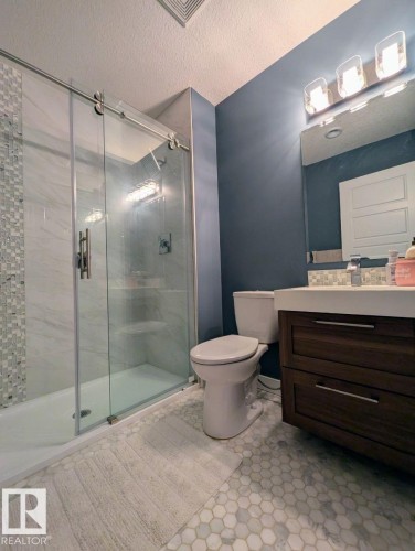 Modern bathroom featuring a frameless glass shower enclosure with a sliding barn door system, hexagon mosaic tile flooring, a dark wood-finish vanity with a white countertop, and a contemporary three-light fixture - 2327 Frezenberg Avenue, Edmonton, AB 