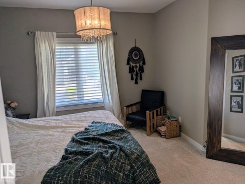 Carpeted bedroom featuring a large window with horizontal blinds, a decorative chandelier, and light gray wall paint - 2327 Frezenberg Avenue, Edmonton, AB 