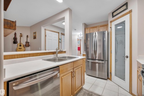Kitchen featuring wood cabinetry, stainless steel appliances including a refrigerator and dishwasher, a double basin sink with a gooseneck faucet, white countertops, and white tile flooring - 207-4807 43A Ave, Leduc, AB 