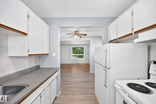 Functional kitchen featuring white cabinetry, a stainless steel sink, and white appliances, with an adjacent room offering wood-finish flooring and a ceiling fan - 355 Northgate Terrace, Edmonton, AB 