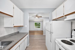 Functional kitchen featuring white cabinetry, a stainless steel sink, and white appliances, with an adjacent room offering wood-finish flooring and a ceiling fan - 