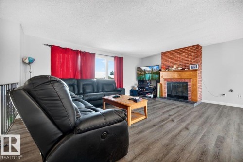 Living room featuring a prominent brick fireplace with a wood mantel, wood-finish flooring, and multiple windows providing natural light - 5215 50 Avenue, Bruderheim, AB - Indoor Photo Showing Living Room With Fireplace
