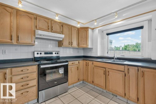Kitchen featuring light wood-finish cabinetry, dark countertops, white tile backsplash, a stainless steel range, and a double-basin stainless steel sink under a window - 16524 99 Street, Edmonton, AB 