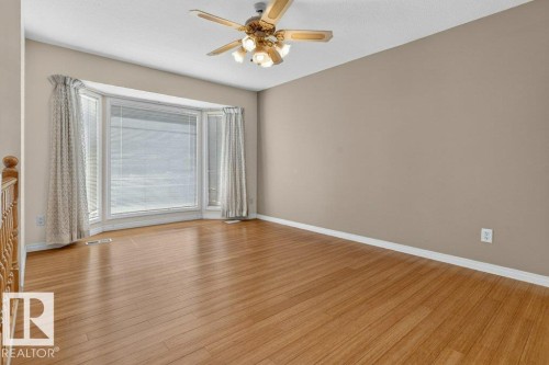 Spacious room featuring a prominent bay window, wood-finish flooring, a ceiling fan with integrated lighting, neutral wall tones, and white trim - 16524 99 Street, Edmonton, AB 