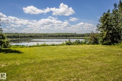 Expansive grassy area bordering a serene body of water, framed by mature trees and lush distant foliage - 