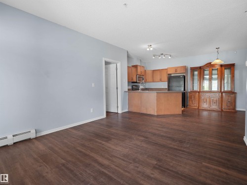 Open-concept living space featuring wood-finish flooring, light blue painted walls, and a kitchen area with wood cabinetry, a built-in oven, and a refrigerator - 106 263 Macewan Road, Edmonton, AB 