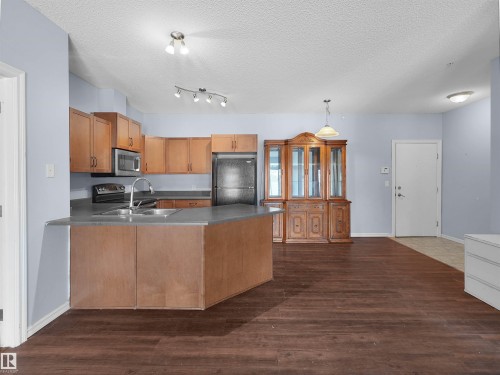 Open-concept kitchen featuring wood-finish cabinetry, stainless steel appliances, and a central island with a double basin sink - 106 263 Macewan Road, Edmonton, AB 