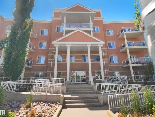 Multi-story building exterior featuring a prominent covered entrance with columns, multiple private balconies with glass railings, and a brick lower facade - 106 263 Macewan Road, Edmonton, AB 