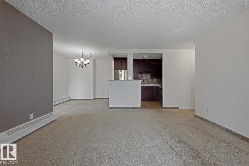 Open concept living area with light-colored carpeting, a partial wall separating the kitchen, and a dining area featuring a chandelier - 505 10145 109 Street, Edmonton, AB 
