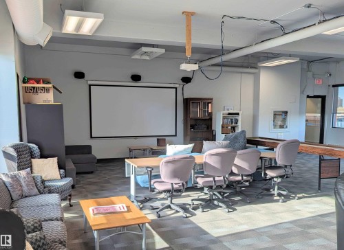 Spacious common area featuring a large projection screen, exposed ceiling ductwork, commercial-style fluorescent lighting, and durable modular carpet flooring - 505 10145 109 Street, Edmonton, AB 