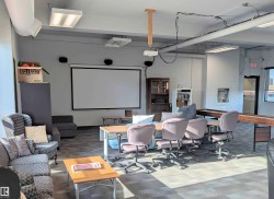 Spacious common area featuring a large projection screen, exposed ceiling ductwork, commercial-style fluorescent lighting, and durable modular carpet flooring - 