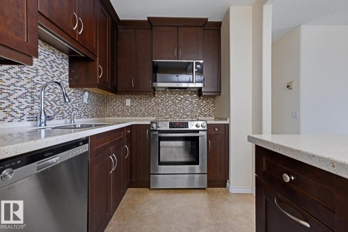 Kitchen featuring dark wood cabinetry, light-toned countertops, a mosaic tile backsplash, and stainless steel appliances including a dishwasher, range, and microwave - 505 10145 109 Street, Edmonton, AB 