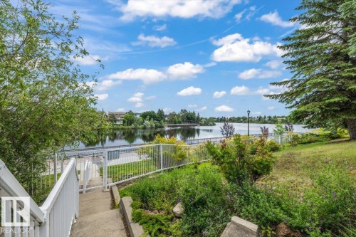 Concrete stairs with white railings leading to a waterfront area featuring a metal fence, surrounding greenery, and a large body of water - 427 15499 Castle Downs Road, Edmonton, AB 