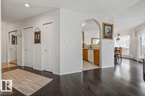 Dark wood-finish flooring extending through the living area, featuring an arched doorway opening to a kitchen with wood cabinetry and a dining area with a large window - 427 15499 Castle Downs Road, Edmonton, AB 