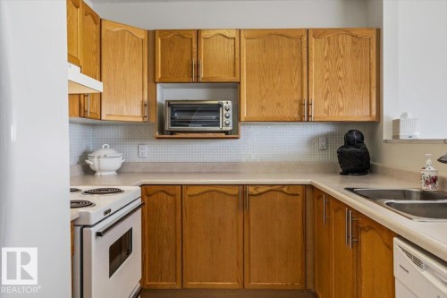 Kitchen featuring light wood-finish cabinetry, a white electric range, a double basin stainless steel sink, and light-colored countertops with a tiled backsplash - 427 15499 Castle Downs Road, Edmonton, AB 