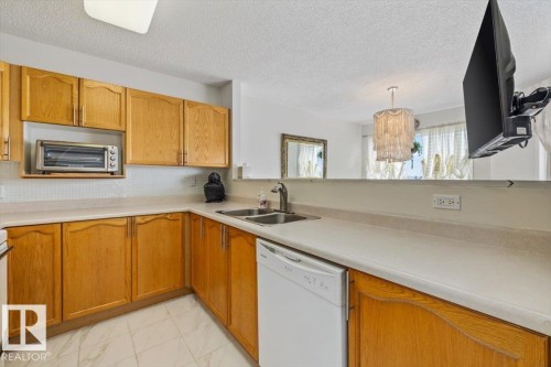 Kitchen featuring wood cabinetry, light-toned countertops, a stainless steel sink with a gooseneck faucet, a white dishwasher, and a white subway tile backsplash - 427 15499 Castle Downs Road, Edmonton, AB 