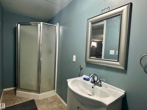 Compact bathroom featuring a corner shower with frosted glass doors, a white pedestal sink with chrome faucet, a framed mirror, and a neutral-toned tile-finish floor - 10619 108 Avenue, Westlock, AB - Indoor Photo Showing Bathroom