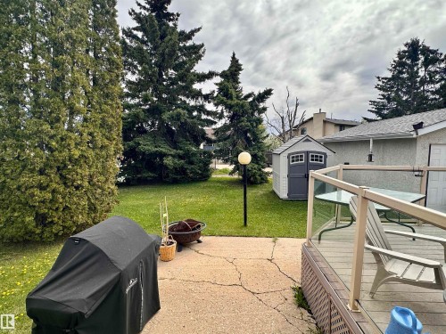 Spacious backyard featuring a wood deck with glass panel railing, a concrete patio, mature evergreen trees, a freestanding shed, and a lawn area - 10619 108 Avenue, Westlock, AB - Outdoor With Deck Patio Veranda