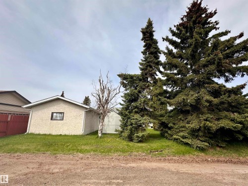 Detached garage with stucco exterior and a pitched shingle roof, accompanied by mature evergreen trees - 10619 108 Avenue, Westlock, AB - Outdoor