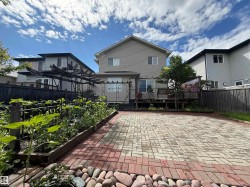 Expansive backyard featuring a large paved patio with a red brick border, raised garden beds with mature plants, and a wooden deck with a sliding glass door - 